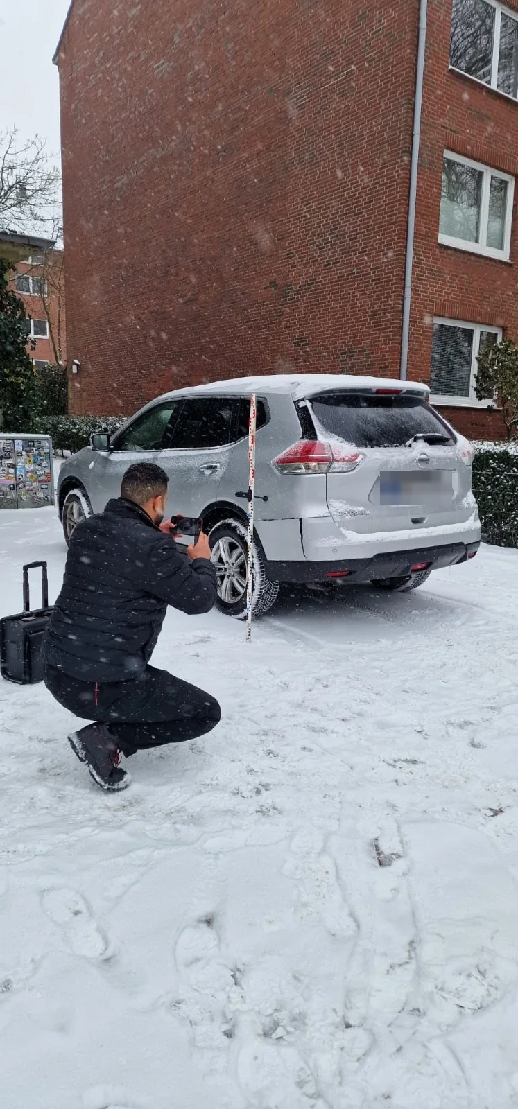 Kfz-Gutachter fotografiert mit dem Smartphone einen Unfallschaden am Heck eines Pkw, daneben eine Messlatte